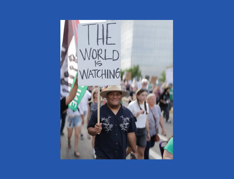 Afa Ah Loo carrying a sign saying, "The World Is Watching"; image from press release.